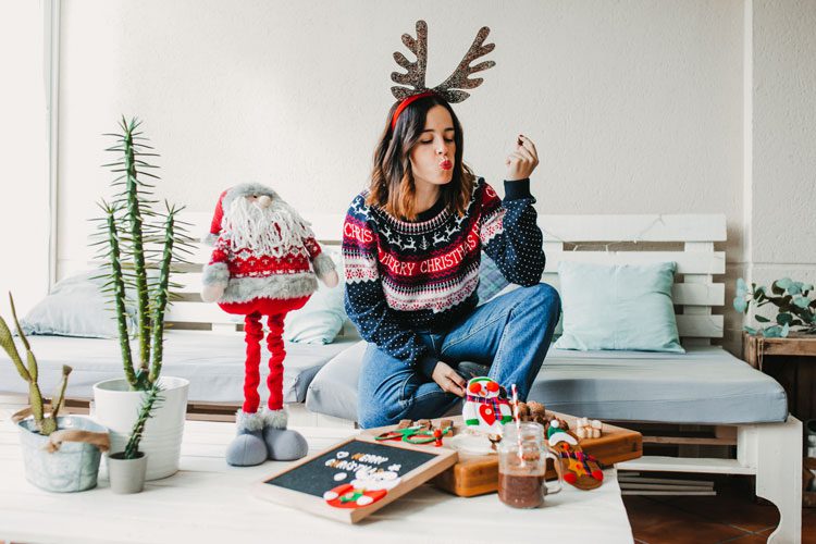 woman in festive sweater and reindeer antlers at home eating sweet treats - the holidays